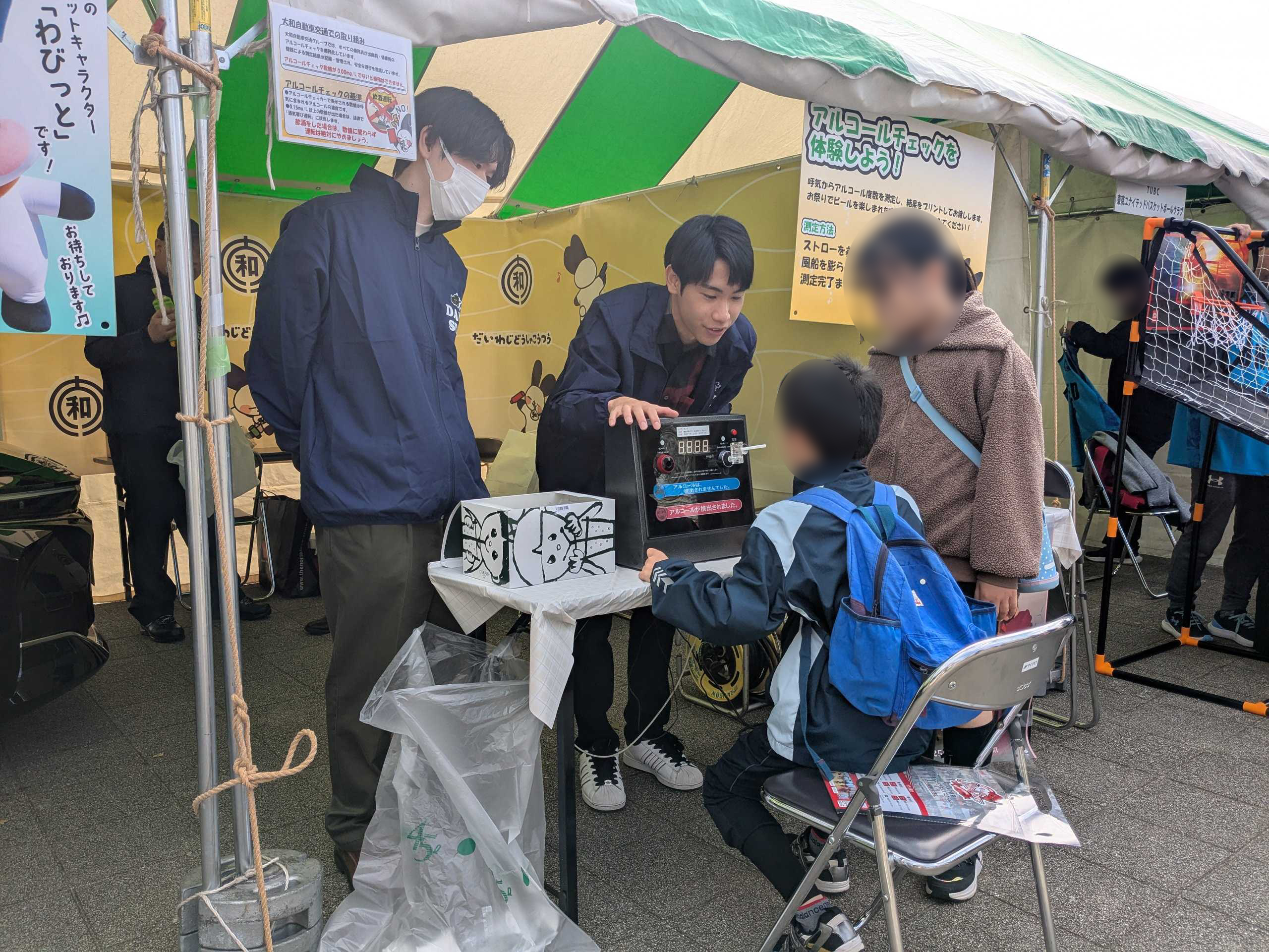 アルコールチェック体験コーナーの写真。スタッフが説明し、参加者は椅子に座って、机の上に設置したアルコールチェッカーにストローで息を吹きかけます。