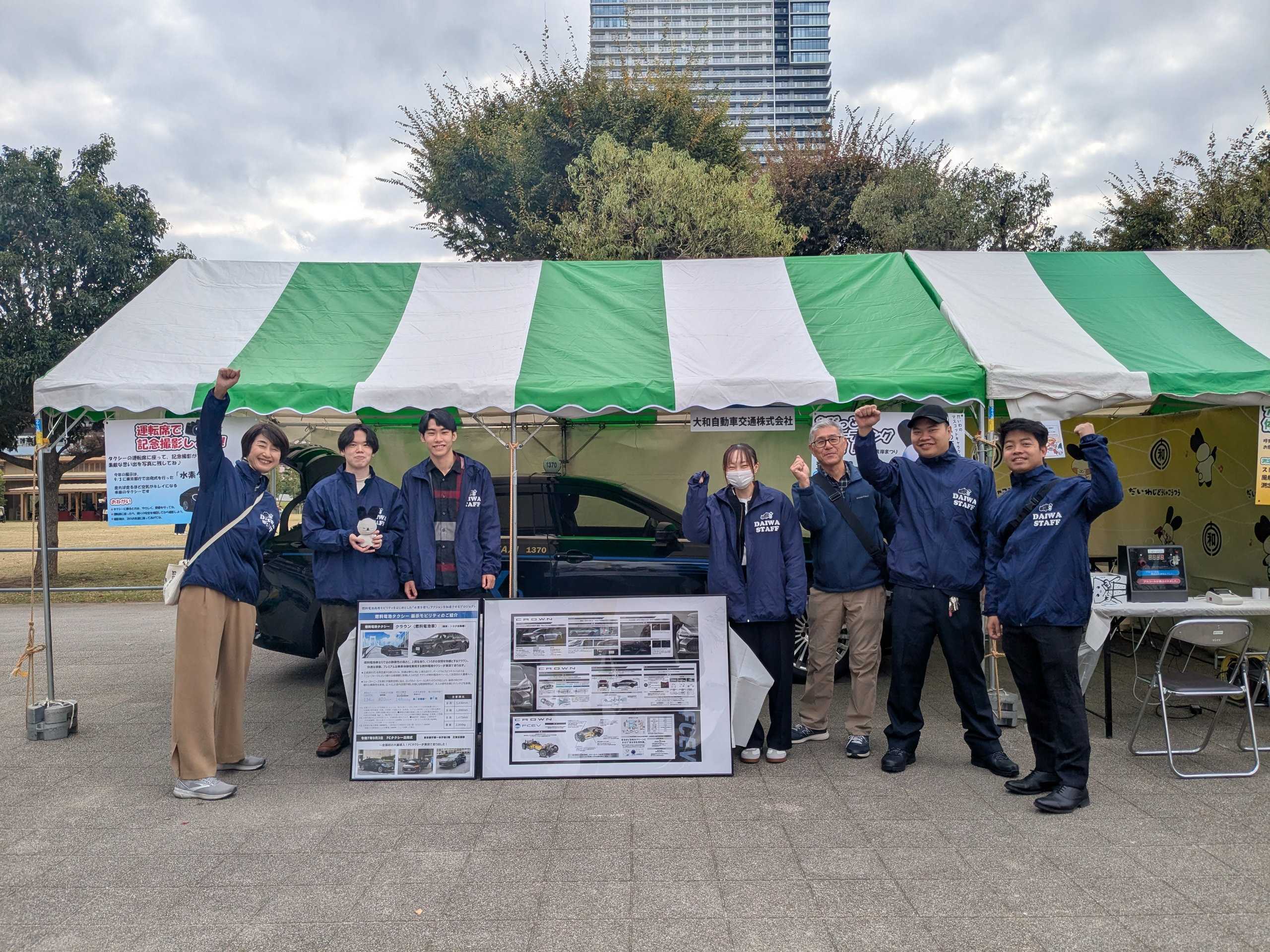 湾岸まつり2025初日のスタッフ集合写真です。水素タクシーのパネルを中心にして７名で撮影しました。