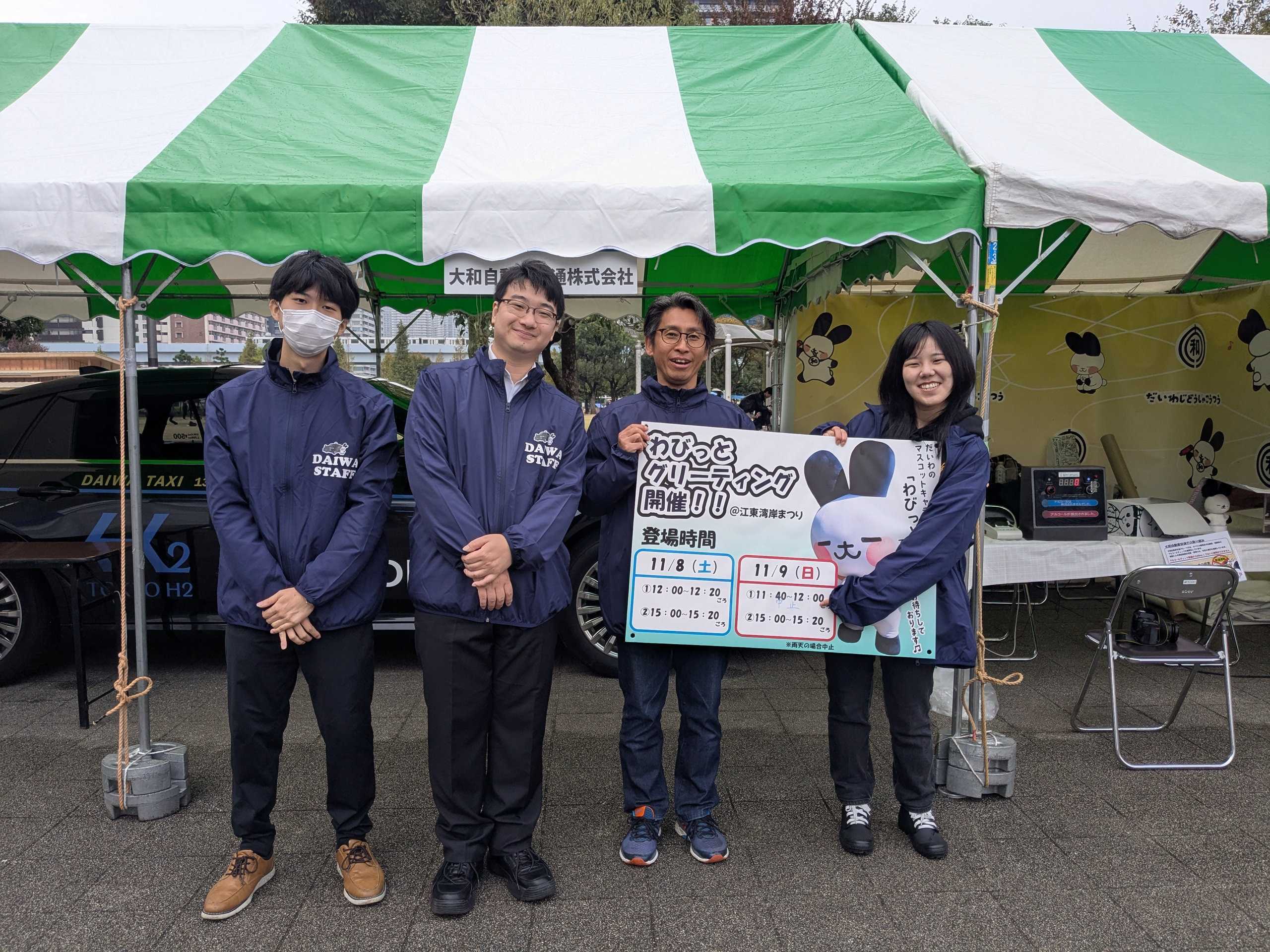 湾岸まつり2025、2日目のスタッフ集合写真です。4名で撮影。そのうち2人は大和自動車交通のマスコットキャラクター「わびっと」着ぐるみグリーティングの案内パネルを持っています。
