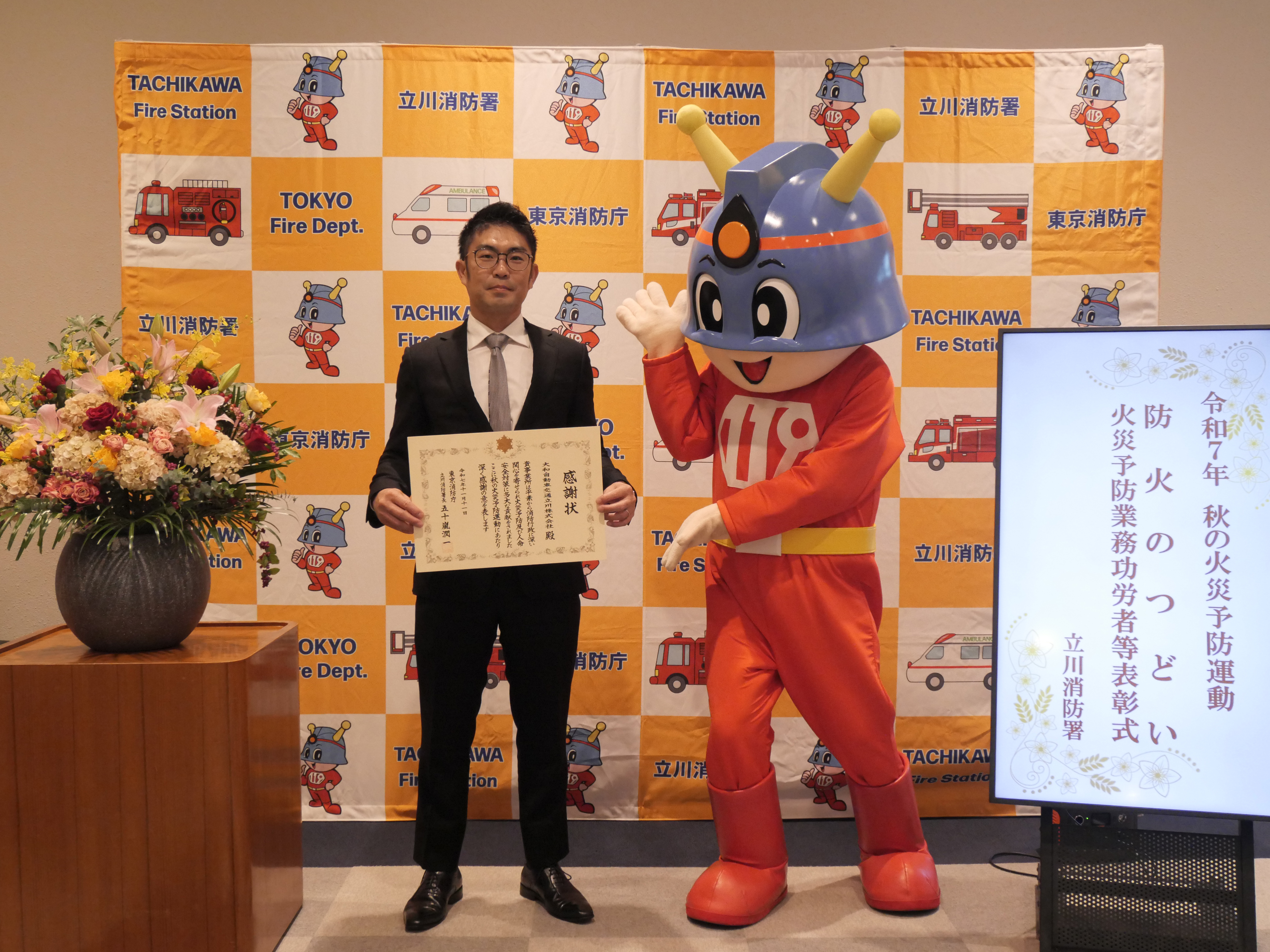 『防火のつどい』終了後、大和自動車交通立川㈱澤田社長が感謝状を持って、東京消防庁マスコットキャラクターの「キュータ」と一緒に記念撮影している写真。