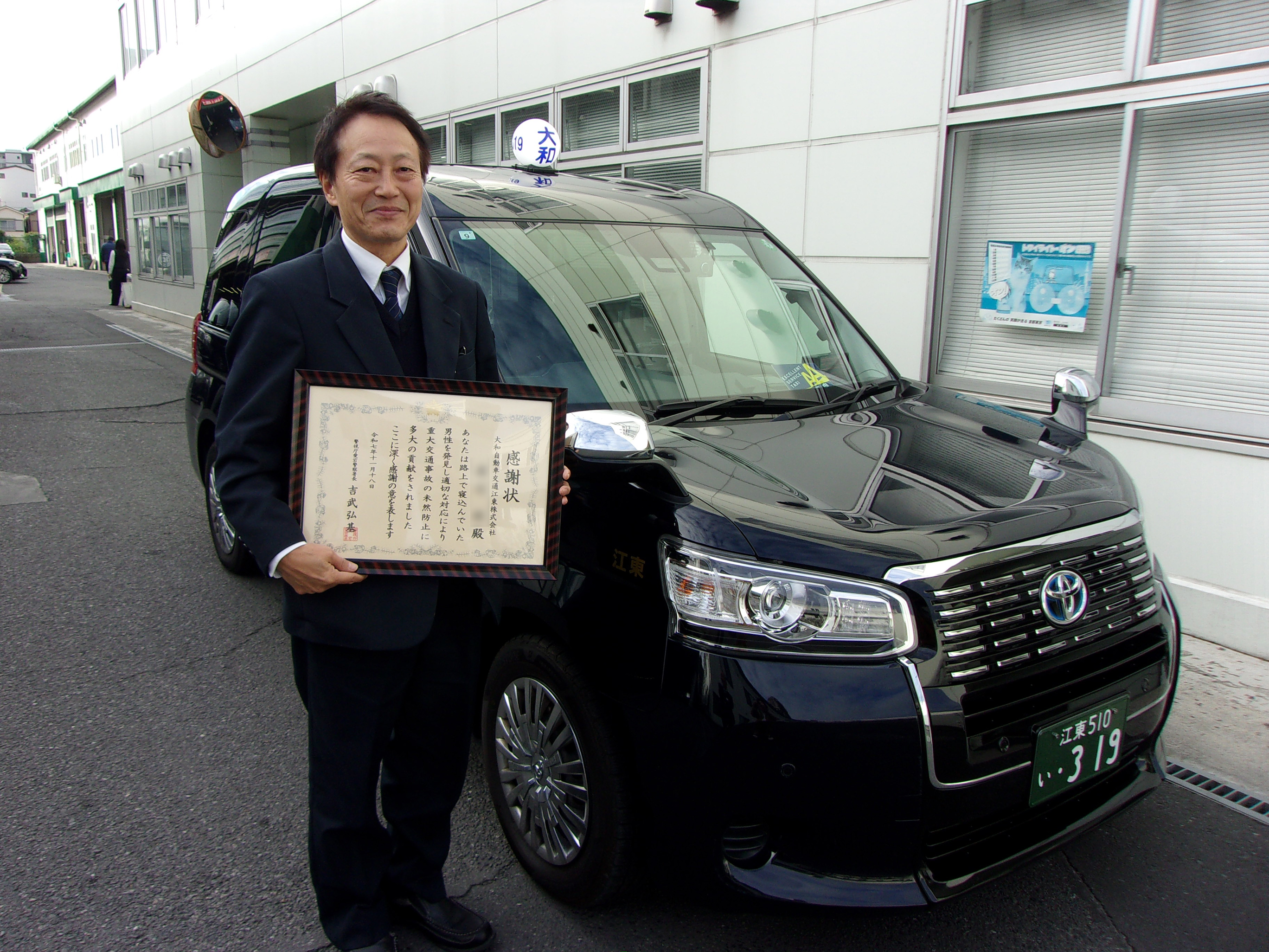 大和自動車交通江東㈱の乗務員が、タクシー車両の前で額に入った感謝状を手に微笑んでいる写真。