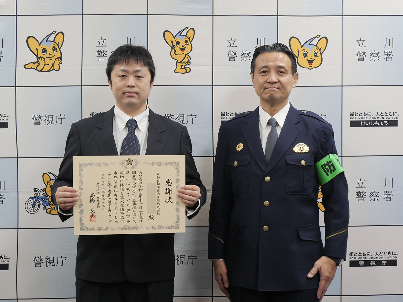 大和自動車交通立川㈱のタクシー乗務員が感謝状を持ち、立川警察署長の方と共に正面を向いている写真。